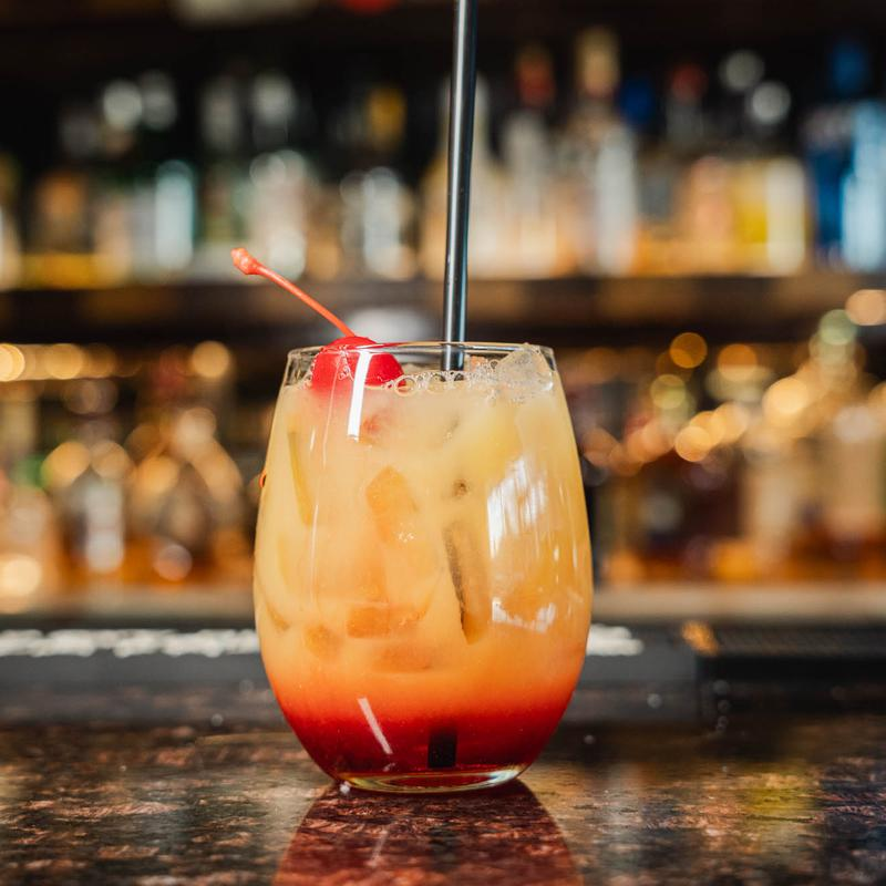 A layered orange cocktail in a rounded glass with ice, a red cherry garnish, and a black straw, set on a dark bar counter.