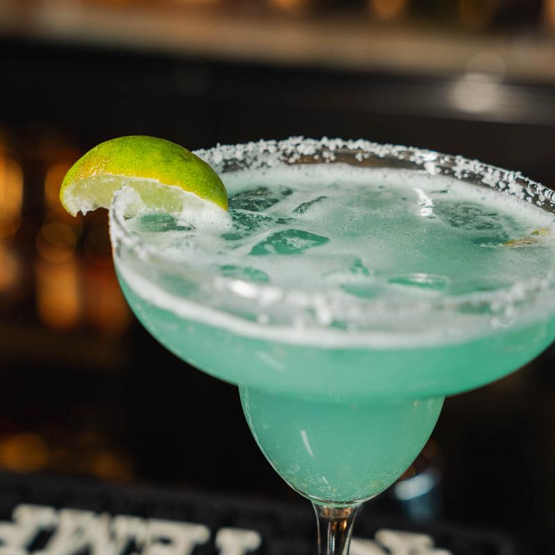 A light blue margarita in a salt-rimmed glass, garnished with a fresh lime wedge on a dark, out-of-focus background.