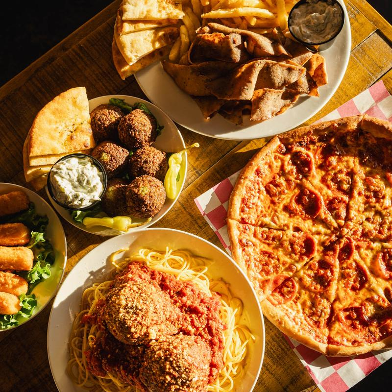 A top-down view of a wooden table featuring various dishes, including pizza, spaghetti with meatballs, pita, and gyros.