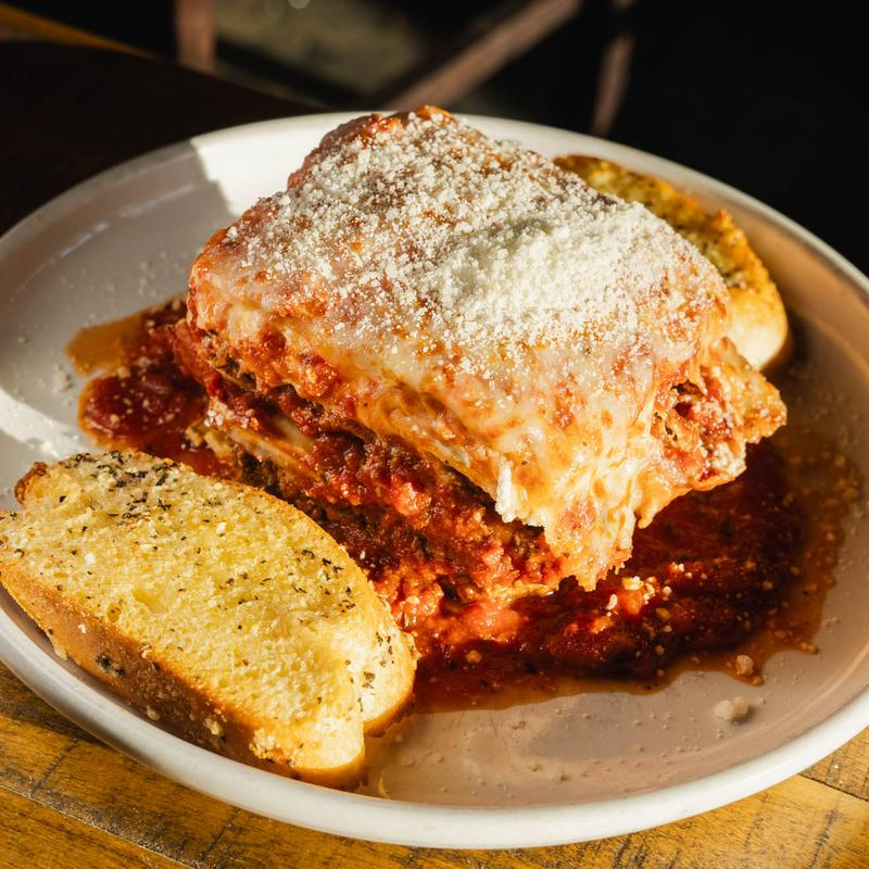A square serving of lasagna topped with melted cheese and parmesan, served with two pieces of toasted garlic bread.