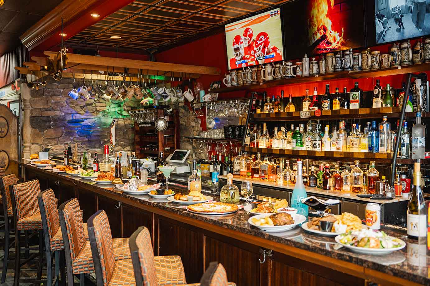 A bar counter lined with several plates of food and stools in a dimly lit, rustic pub with shelves of liquor bottles.
