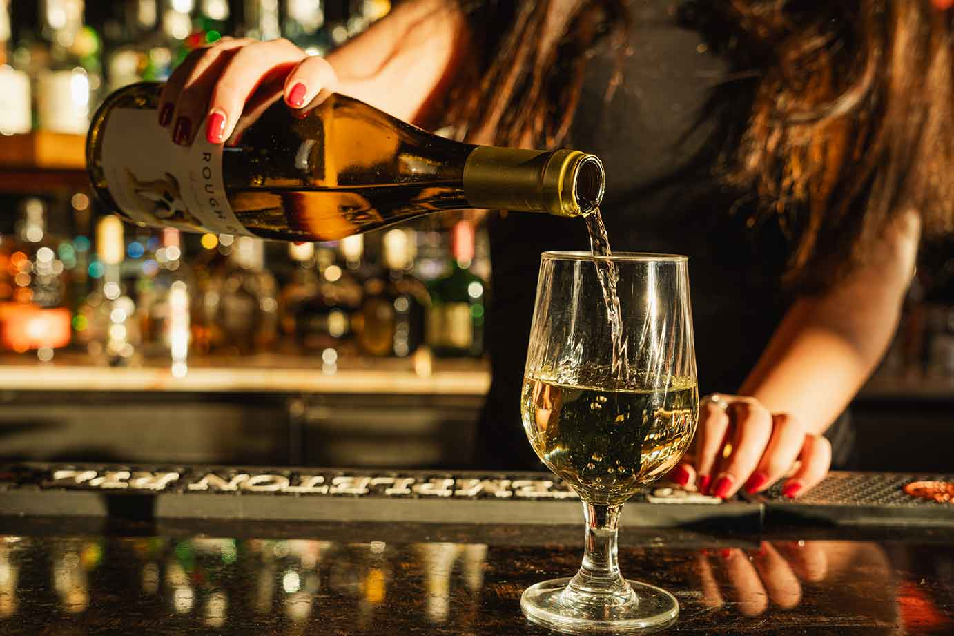A person pours white wine from a bottle into a stemless glass on a bar counter with a blurred background of liquor bottles.