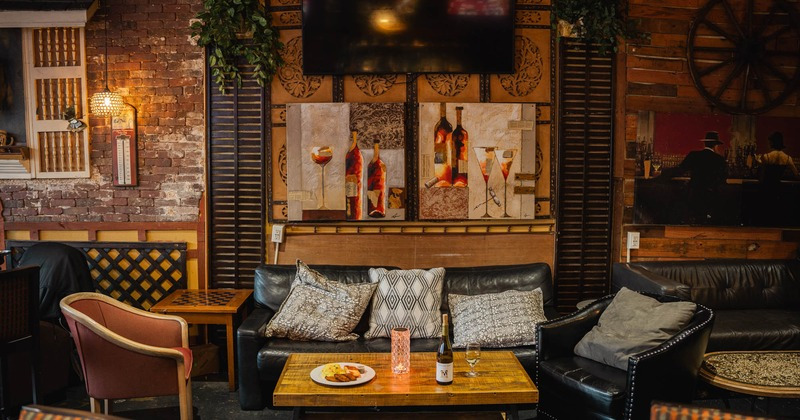 A cozy restaurant seating area with black leather sofas, armchairs, a wooden coffee table, and wine-themed wall art.