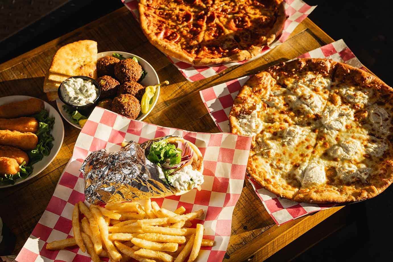 Top-down view of a wooden table featuring two pizzas, a gyro with fries, falafel, and bread sticks on checkered paper.
