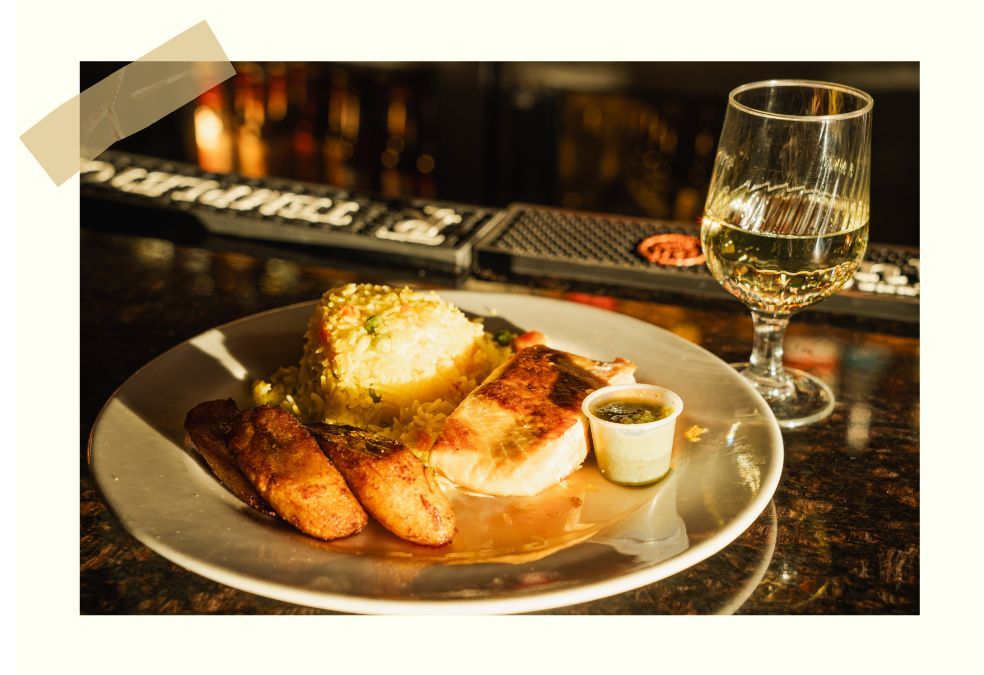 A dinner plate featuring grilled fish, fried plantains, and a scoop of rice next to a glass of white wine on a bar counter.