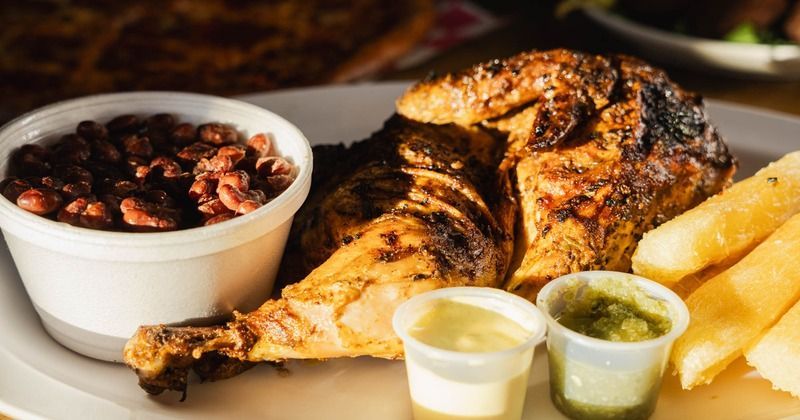 A roasted chicken platter served with black beans, yuca fries, and small containers of yellow and green sauces.