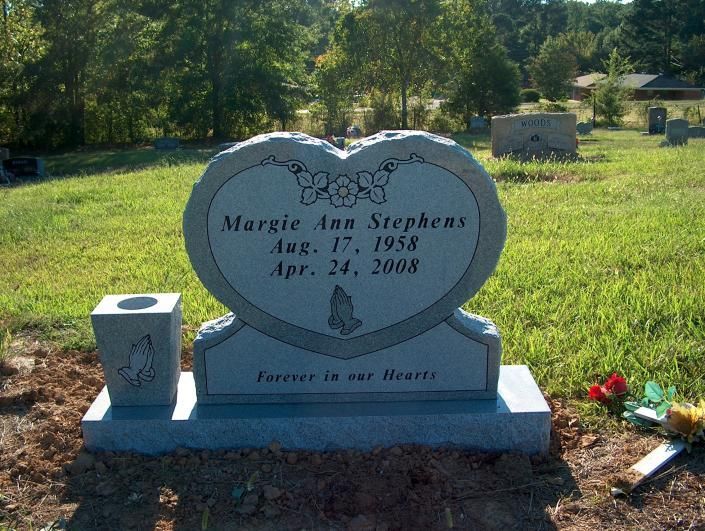 Heart-shaped gravestone in a green cemetery with flowers and a small memorial plaque.