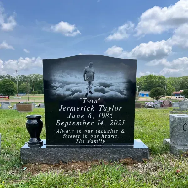 Black gravestone with a portrait, white text, and a vase in a grassy cemetery under a cloudy sky