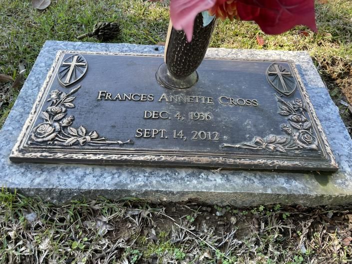 Bronze memorial plaque with engraved cross and floral border, topped by red flowers in a grassy cemetery.