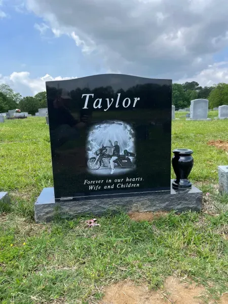 Black gravestone engraved “Taylor” with a photo and memorial text, set in a grassy cemetery.