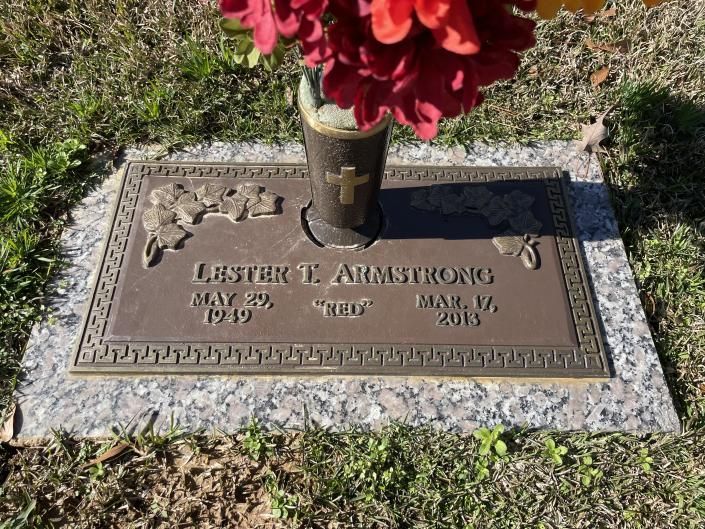 Gravestone in grass with red flowers at the top, memorial plaque reading Lester L. Armstrong.