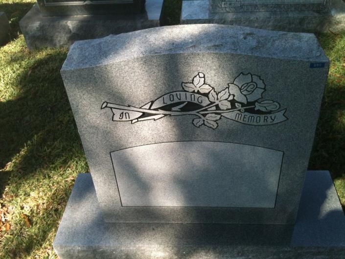Gray memorial headstone with carved flowers and a ribbon, partly shaded by sunlight on grass.