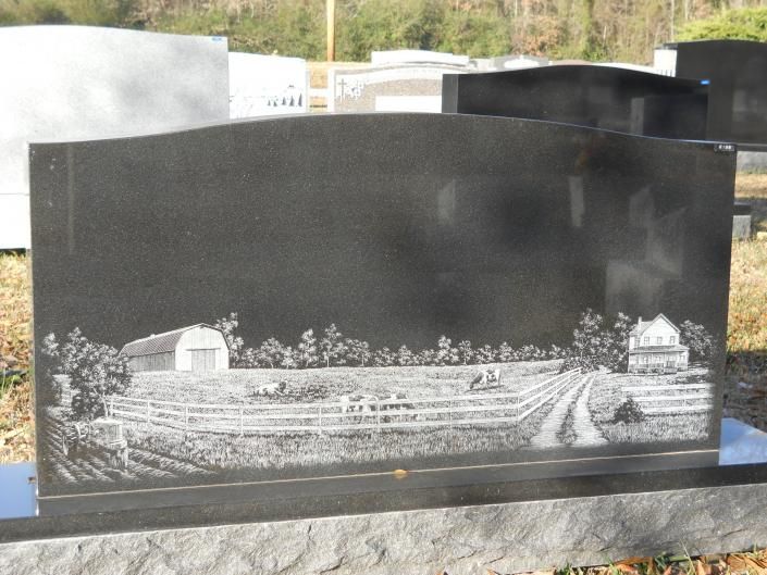 Black granite gravestone engraved with a rural landscape scene of fields, trees, and a farm