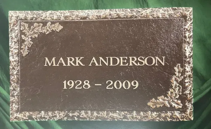Gravestone plaque reading “MARK ANDERSON, 1928–2009” with ornate floral border.