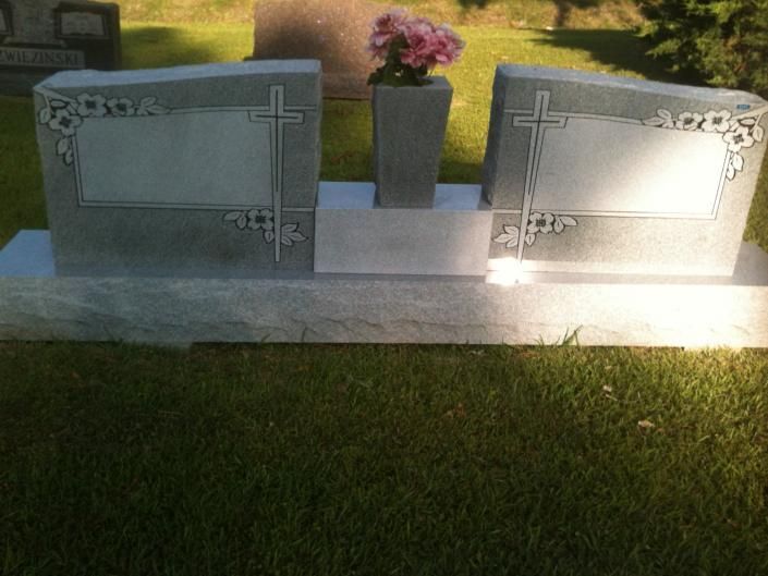 Two upright gravestones with floral etchings and a flower vase in the center, set on a lawn at sunset