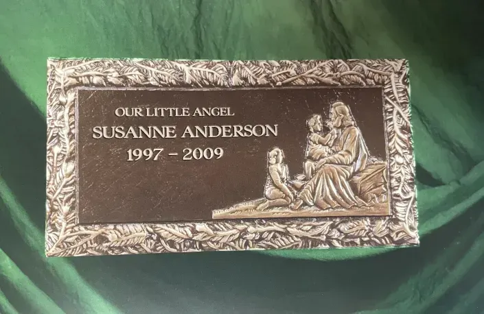 Memorial plaque with angel relief and inscription “Our little angel, Susanne Anderson, 1997–2009” on green cloth