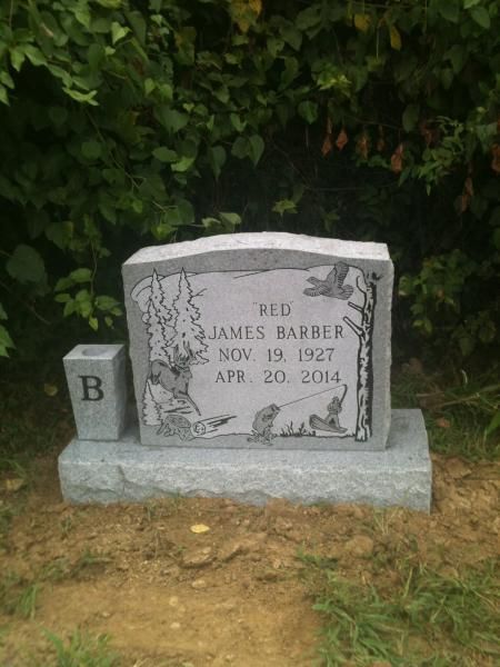 Gravestone with “B” marker in front, set against greenery on a dirt patch.