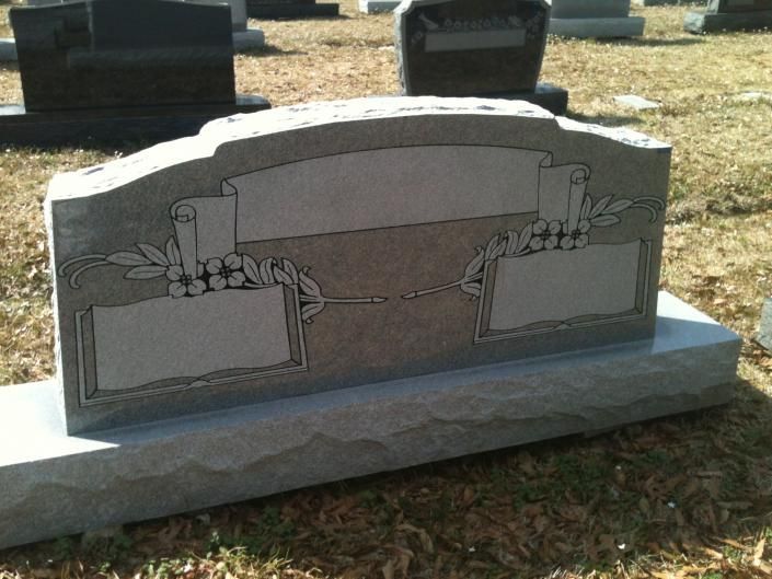 Small gray cemetery headstone with engraved scroll and flowers on a stone base, outdoors on grass
