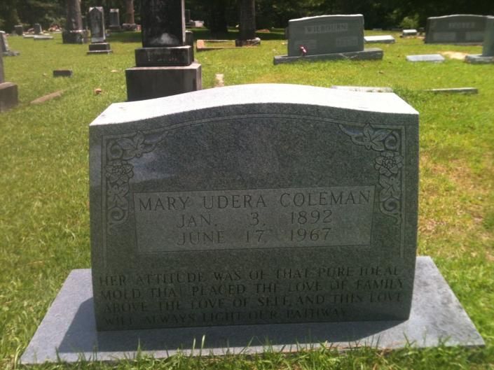 Gray gravestone in a cemetery with engraved name and dates, surrounded by other headstones on green grass