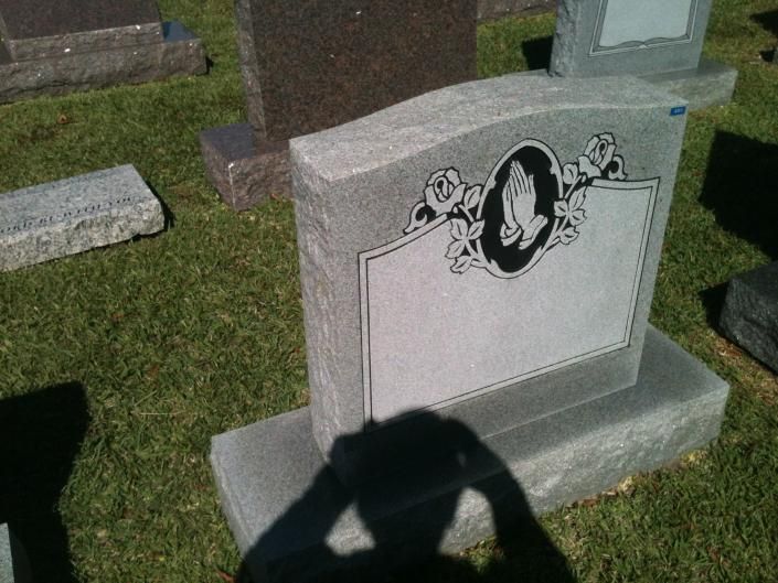 Gray gravestone with decorative plaque in a cemetery, seen in sunlight with photographer’s shadow in foreground