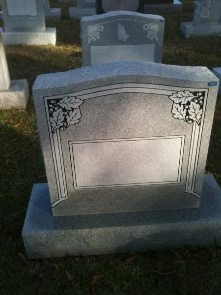 Gray engraved gravestone in a cemetery on grass, with another headstone blurred behind it.