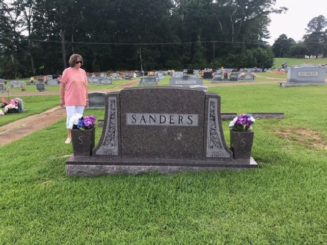 Person in pink stands by a Sanders gravestone decorated with flowers in a cemetery.
