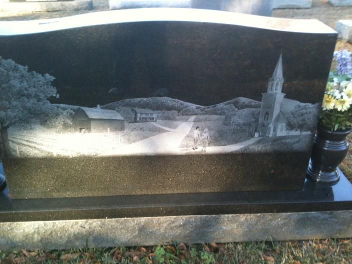 Black engraved memorial stone with a village scene and church steeple, set on a granite base outdoors
