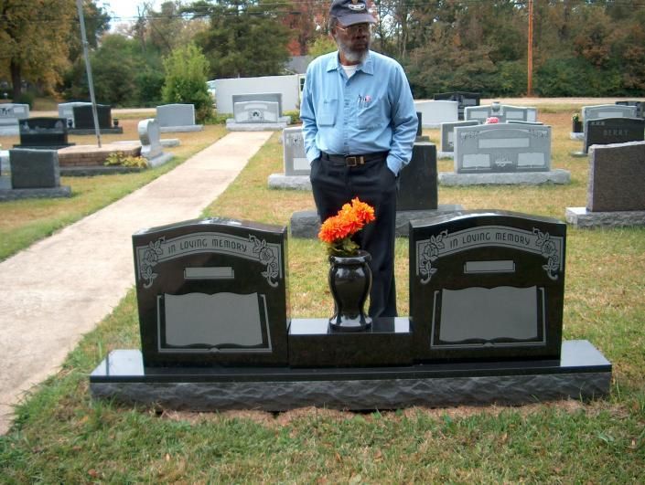 Man standing behind a black double grave marker with flowers in a cemetery