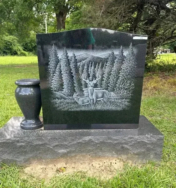 Black granite memorial stone with engraved forest scene, beside a small urn-like vase on a stone base outdoors
