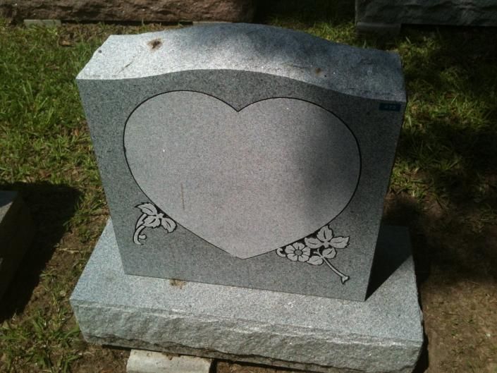 Gray heart-shaped gravestone with floral carvings on a stone base outdoors.