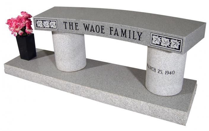 Gray memorial bench with engraved family name, two white supports, and a small pink flower vase