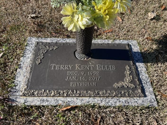 Gravestone with engraved name and dates, topped by yellow flowers in a cemetery.