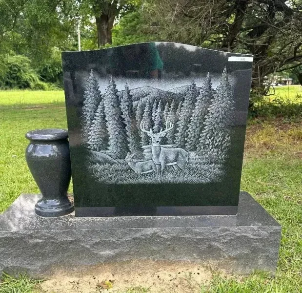 Black granite memorial stone with engraved deer and trees, set on a grassy lawn beside a dark vase.