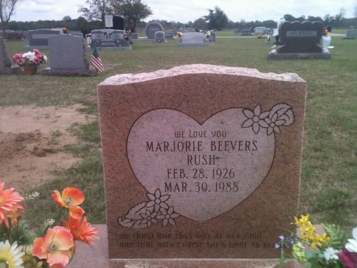 Pink heart-shaped gravestone in a cemetery with engraved names and dates, surrounded by flowers