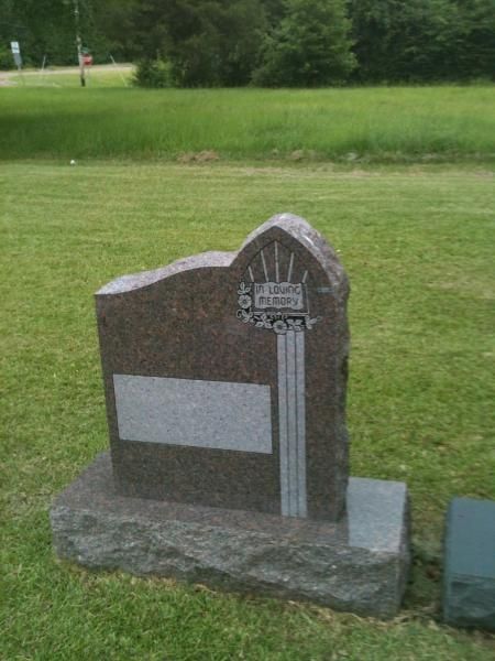 Brown memorial headstone with a white plaque in a grassy cemetery, with a small decorative emblem at the top.