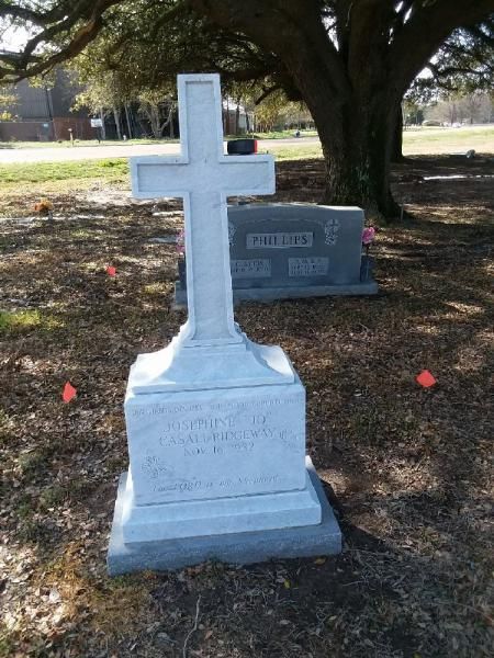 White cross gravestone in a cemetery under trees, with a smaller gray headstone behind it.