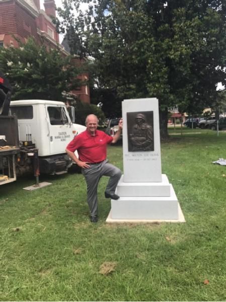Man in red shirt posing beside a white monument-style sculpture on a grassy lawn, with a truck and trees behind.