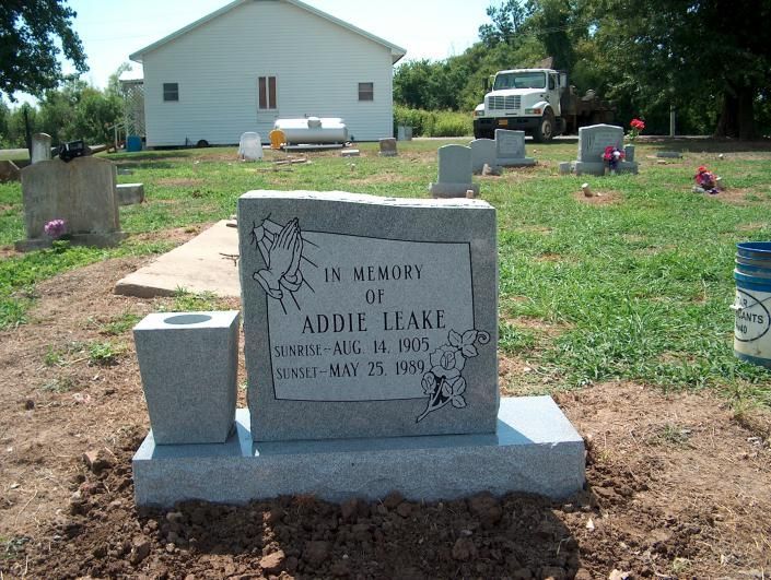 Gravestone in a cemetery, with a house and truck in the background.