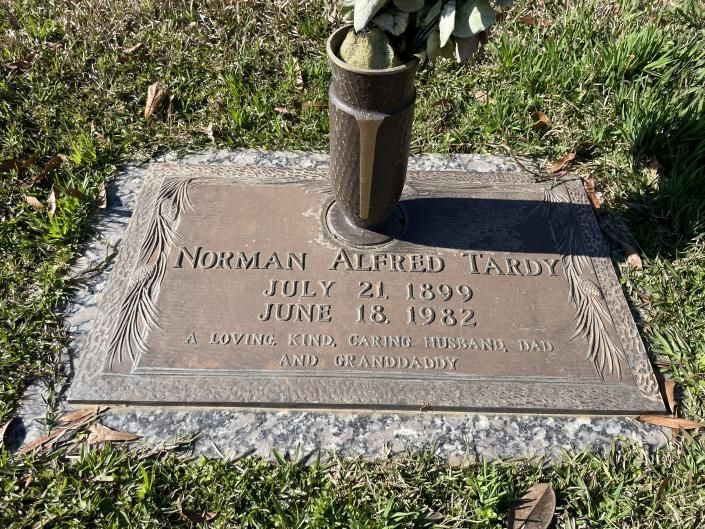 Gravestone plaque with engraved name and dates, topped by a vase of flowers in grass and sunlight