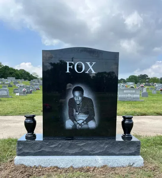 Black gravestone with “FOX” and a portrait, set in a cemetery under a cloudy sky