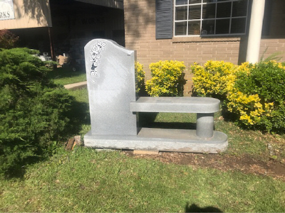 Gray memorial bench monument beside a building, with yellow shrubs and grass