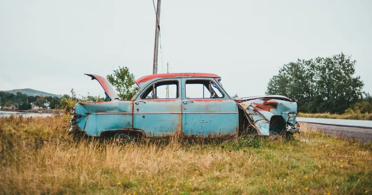 Rusty blue vintage car wreck in a grassy field beside a road under an overcast sky
