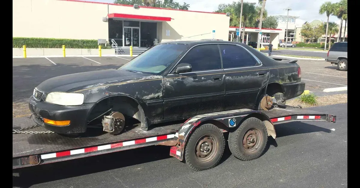 Black sedan strapped on a dual-axle trailer in a parking lot, with a building in the background
