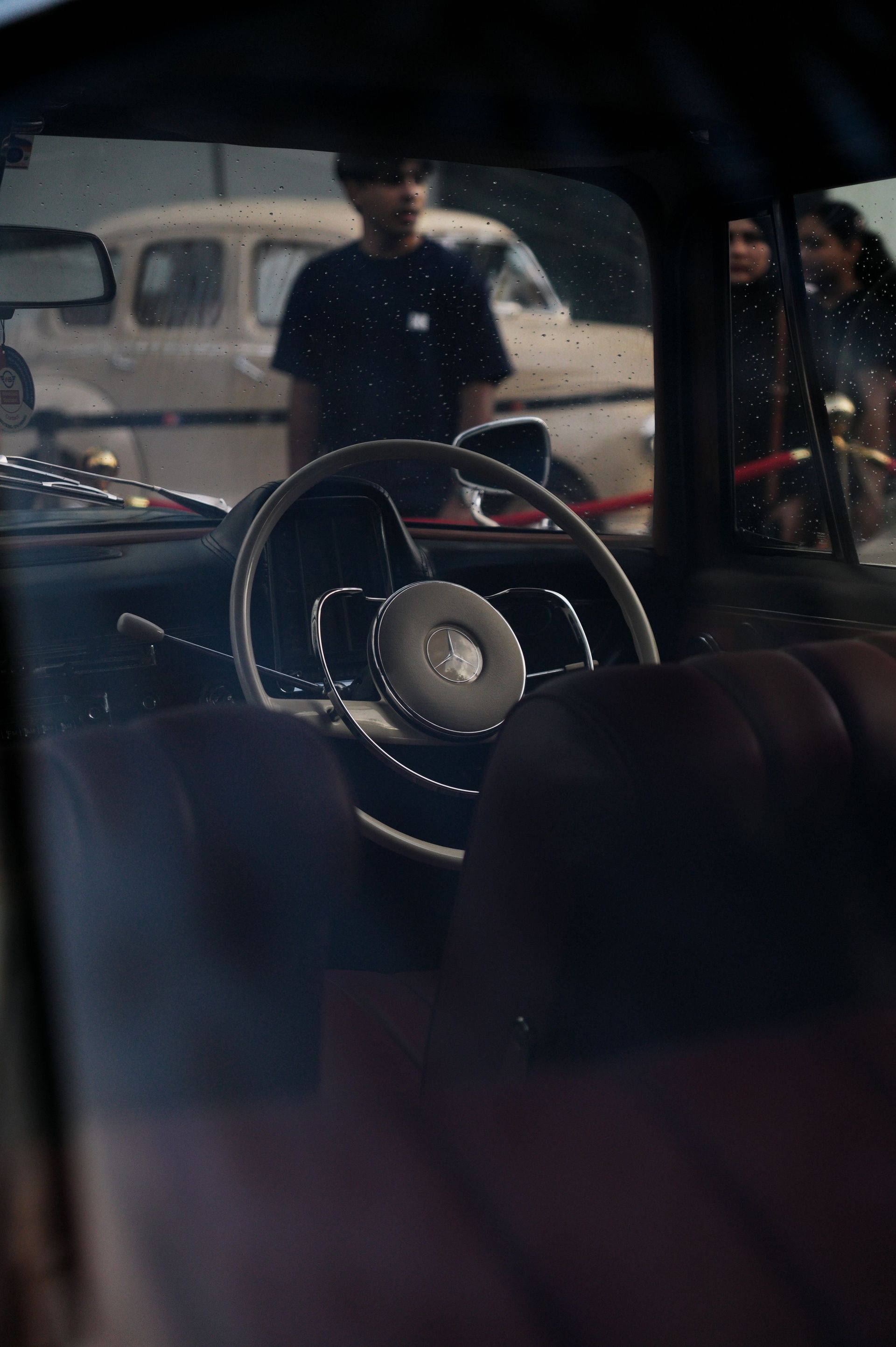 Car interior view with steering wheel, parked cars, and two people standing outside.