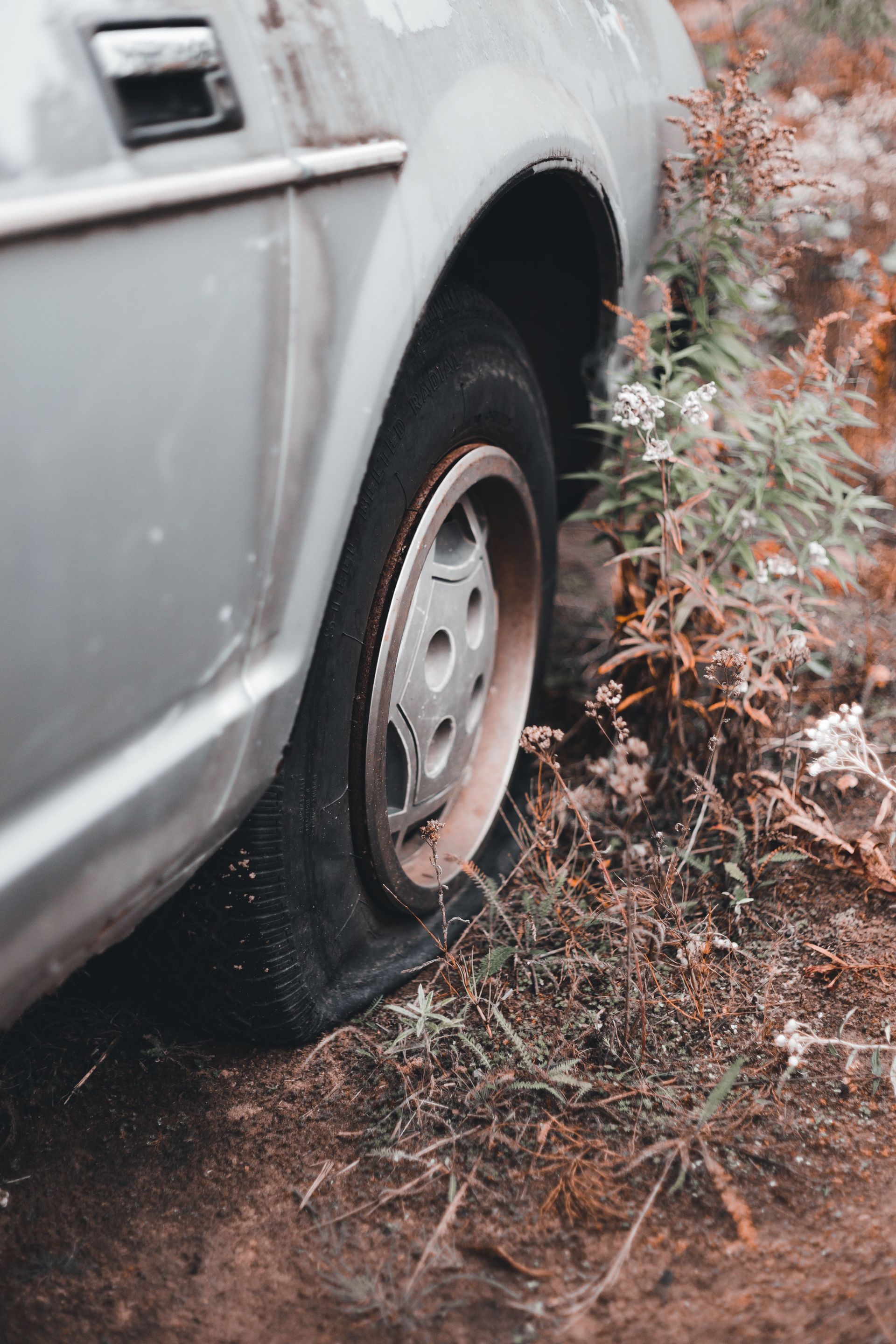 Silver car stuck in muddy grass beside bushes, with a flat front tire