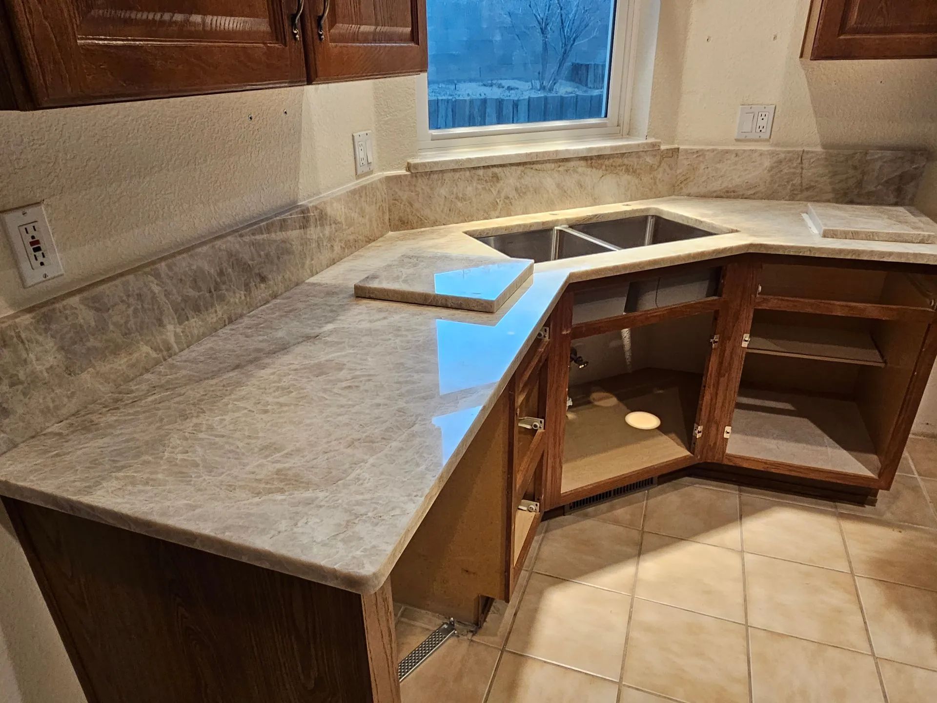 Kitchen corner with installed granite countertop, open cabinet doors, and unfinished sink area near a window.