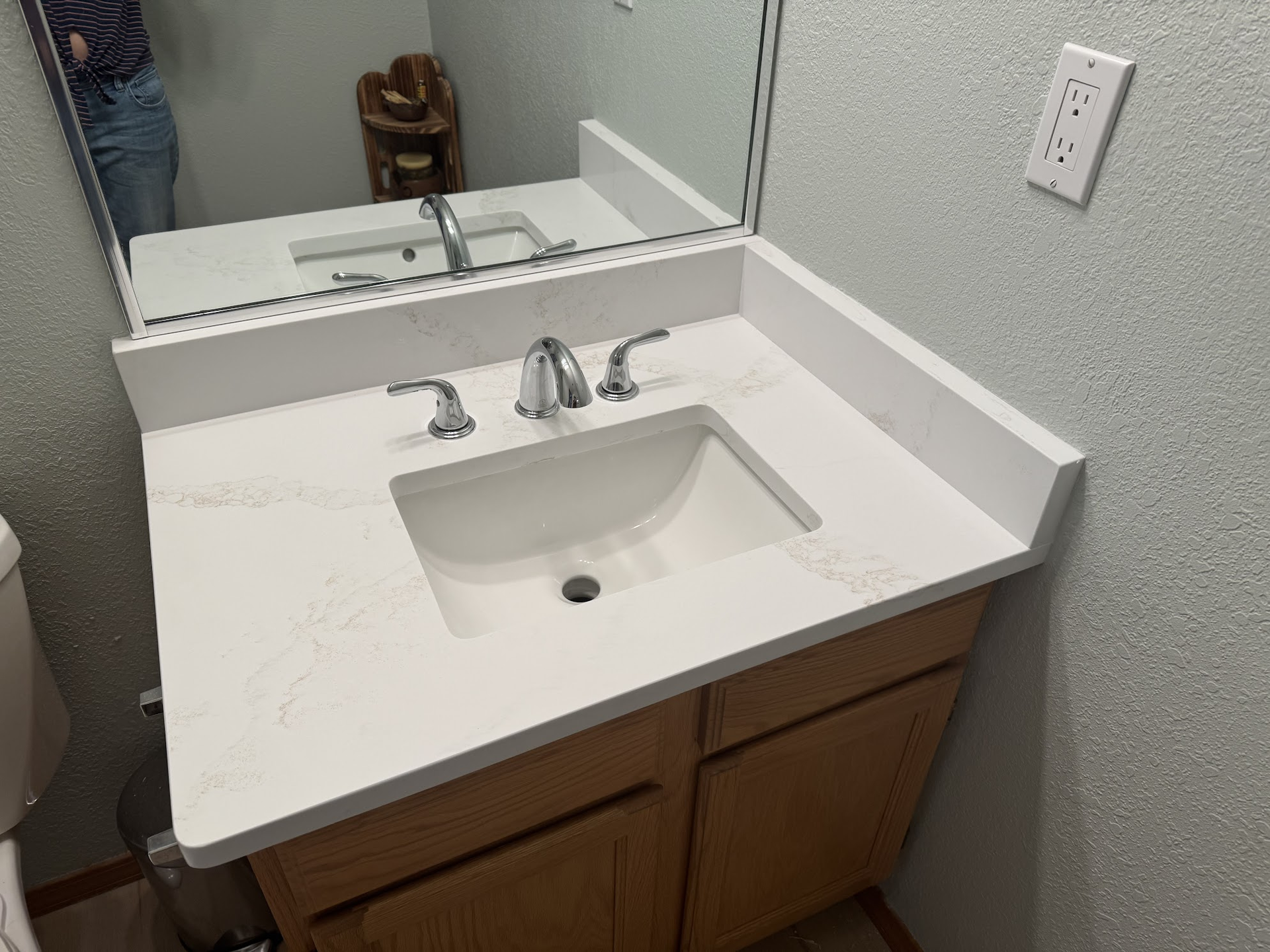 Bathroom vanity with white countertop, sink, and silver faucet. Light brown cabinet.
