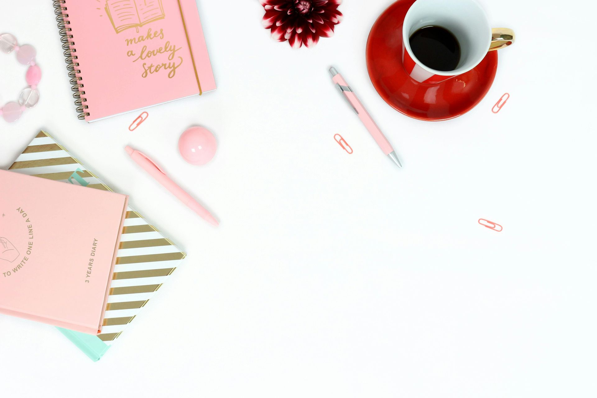 A white table topped with pink notebooks , pens , a cup of coffee and a bracelet.