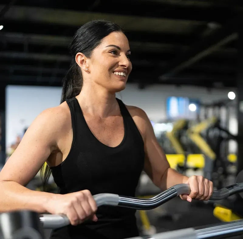 Woman using a gym machine, smiling in a black tank top, with blurred equipment in the background