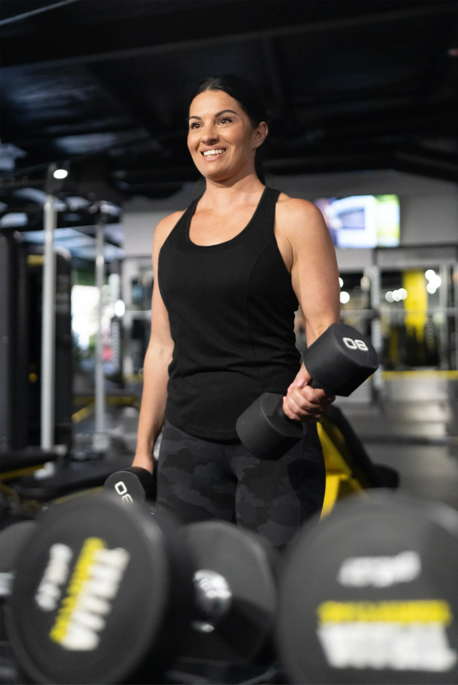 Woman lifting dumbbells in a gym, smiling in a black tank top amid blurred weights.